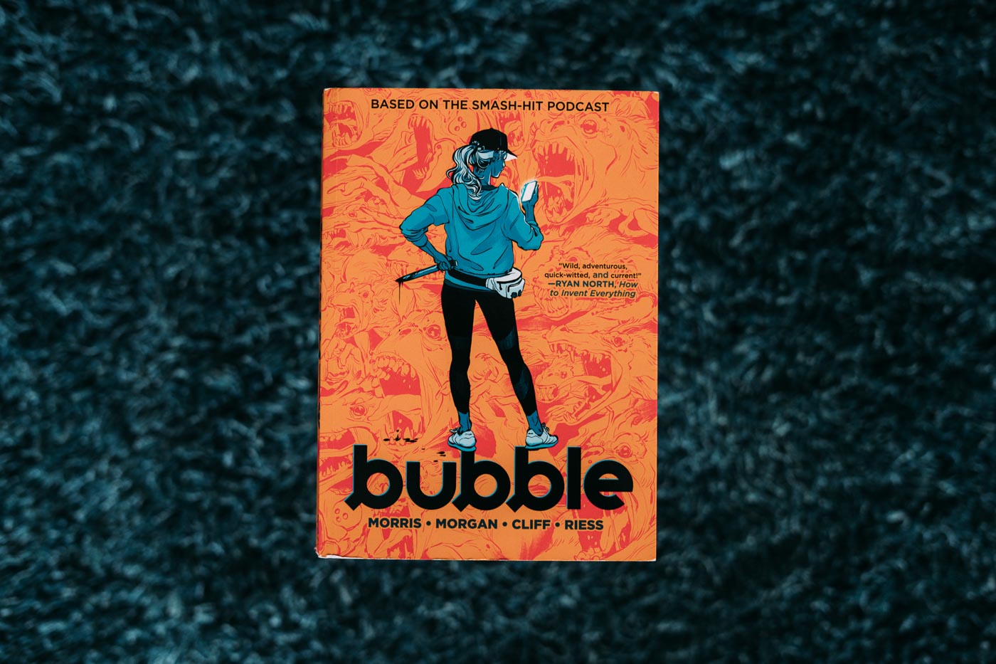 Ett seriealbumsomslag. En kvinna i träningskläder och utrustad med magväska står och läser i sin telefon. Bakom ryggen håller hon en blodig kniv. Titeln lyder Bubble.