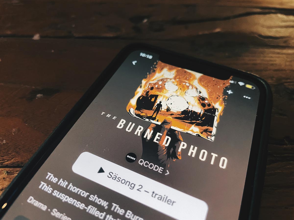 Närbild av en iPhone som spelar trailern av säsong två av podcasten The Burned Photo. Omslagsbilden är just på ett brinnande foto. Fotot föreställer en person i siluett som betraktar ett brinnande hus.